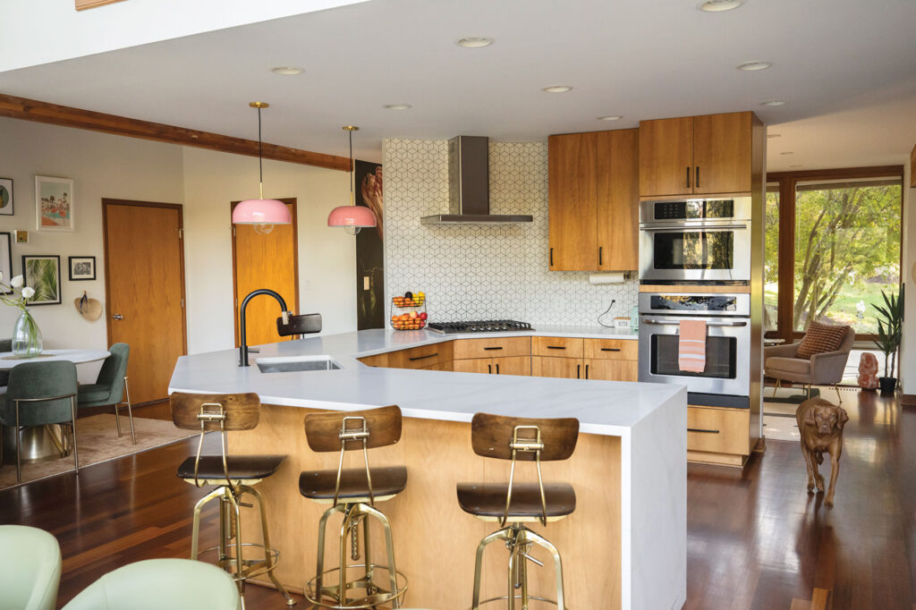 Interior designer Ana Tavares’s kitchen featuring quartz countertop, bright pink and brass pendant lights, a monochromatic backsplash and wood slab cabinets.