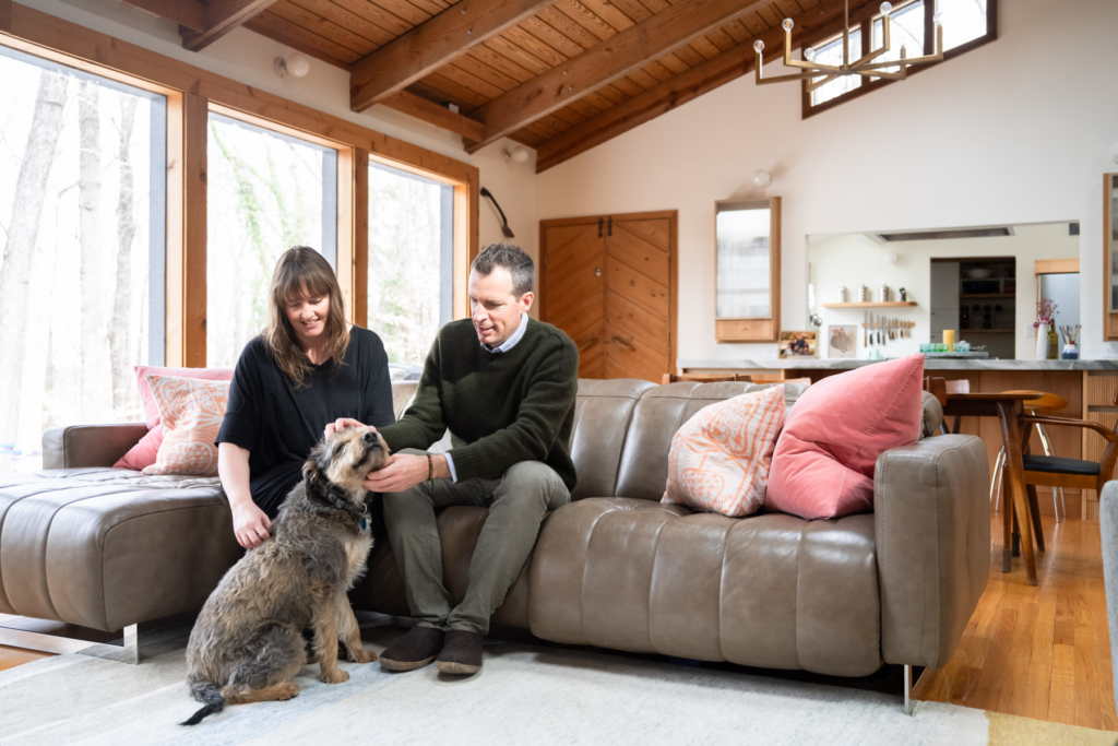A man and a woman sit on a couch while they lovingly pet a dog.