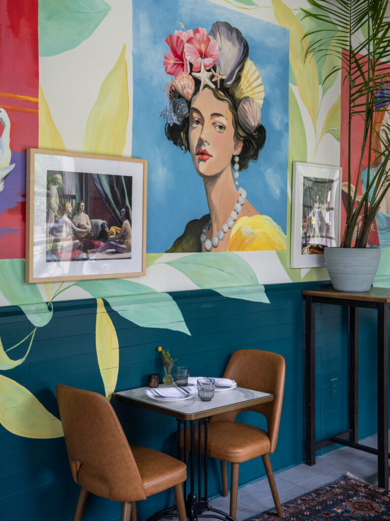 Two-top table with tan leather chairs beneath a mural of a woman wearing seashells and hibiscus in her hair, flanked by framed prints on a teal and cream wall.
