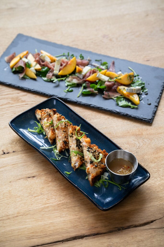 Sesame shrimp toast triangles with scallions and dipping sauce on a blue plate, with a slate board of peaches, prosciutto, cheese and greens behind.