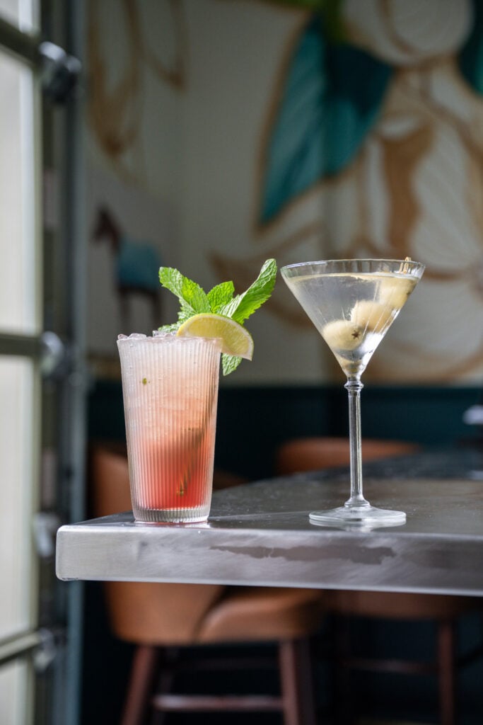 Pink raspberry nojito in a ribbed glass garnished with mint and lime beside a martini with olives on a marble table
