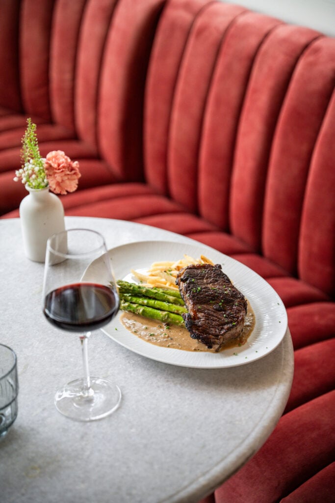 Steak frites with asparagus and pan sauce on a white plate beside a glass of red wine on a marble table in front of a red velvet banquette