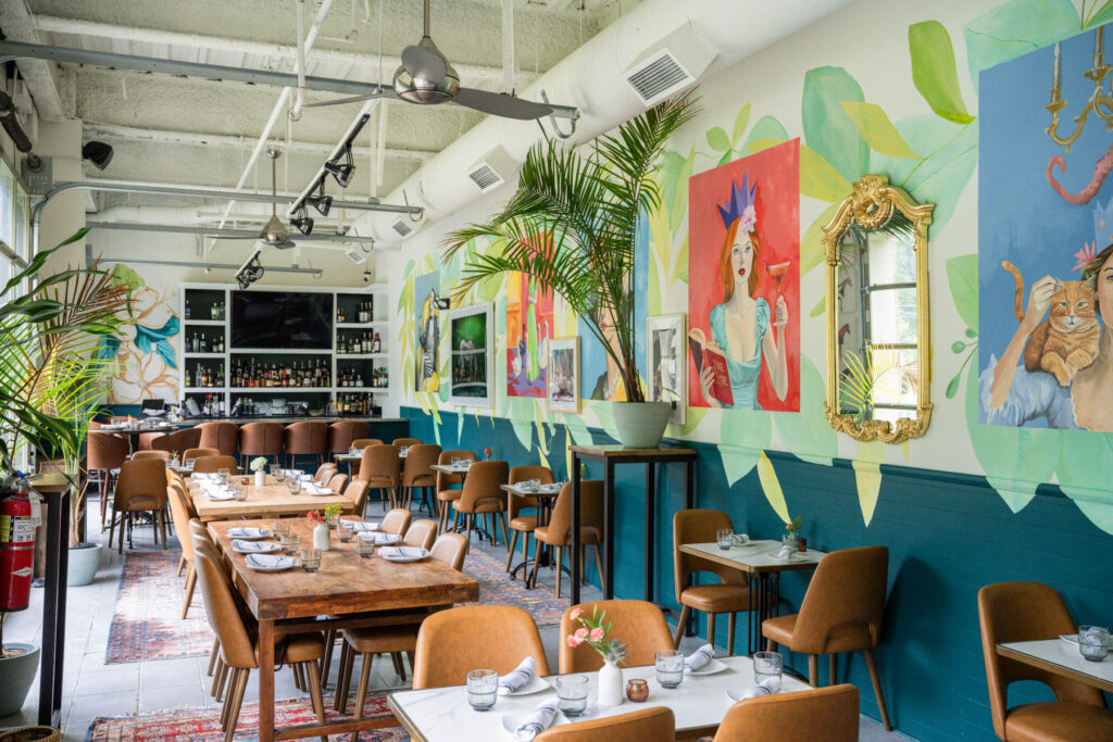 Dining room with tan leather chairs, wood and marble tables, and a wall of hand-painted murals of women, tropical leaves and a gilded mirror, with a bar at the far end