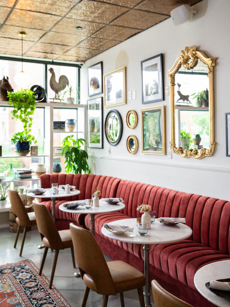 Dining room with a long channel-tufted rose velvet banquette, round marble cafe tables, a gallery wall of framed art and gilded mirrors, and a pressed tin ceiling